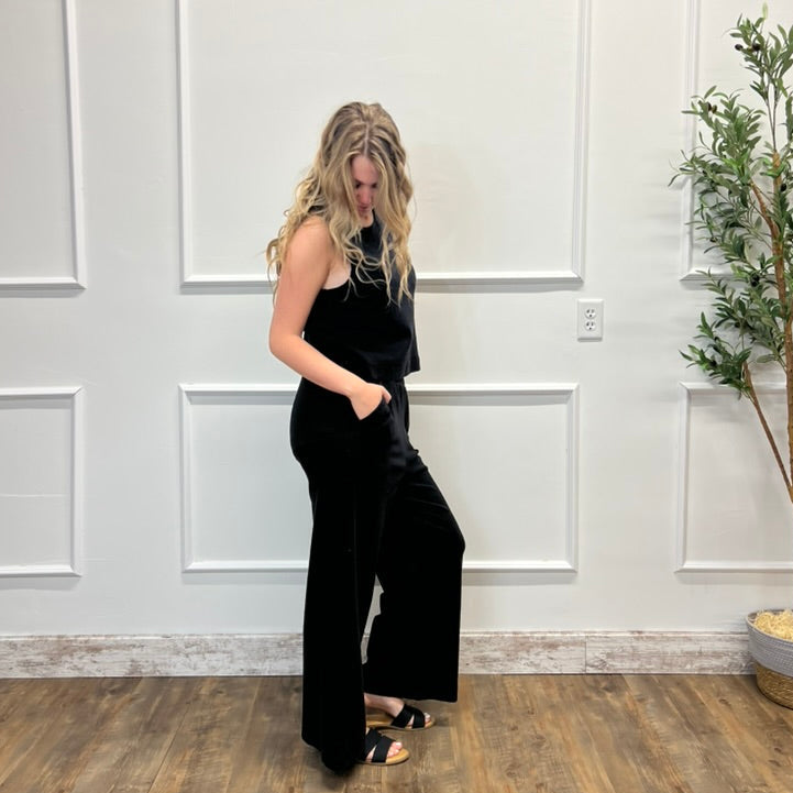 Woman in a black jumpsuit standing in a room with white paneled walls and a plant.