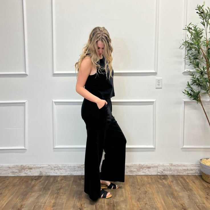 Woman in a black jumpsuit standing in a room with white paneled walls and a plant.