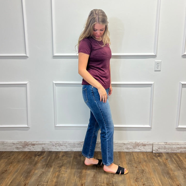 A woman modeling a short-sleeve crewneck t-shirt in heathered plum purple and blue jeans, standing in a room with white paneled walls and a wood floor.