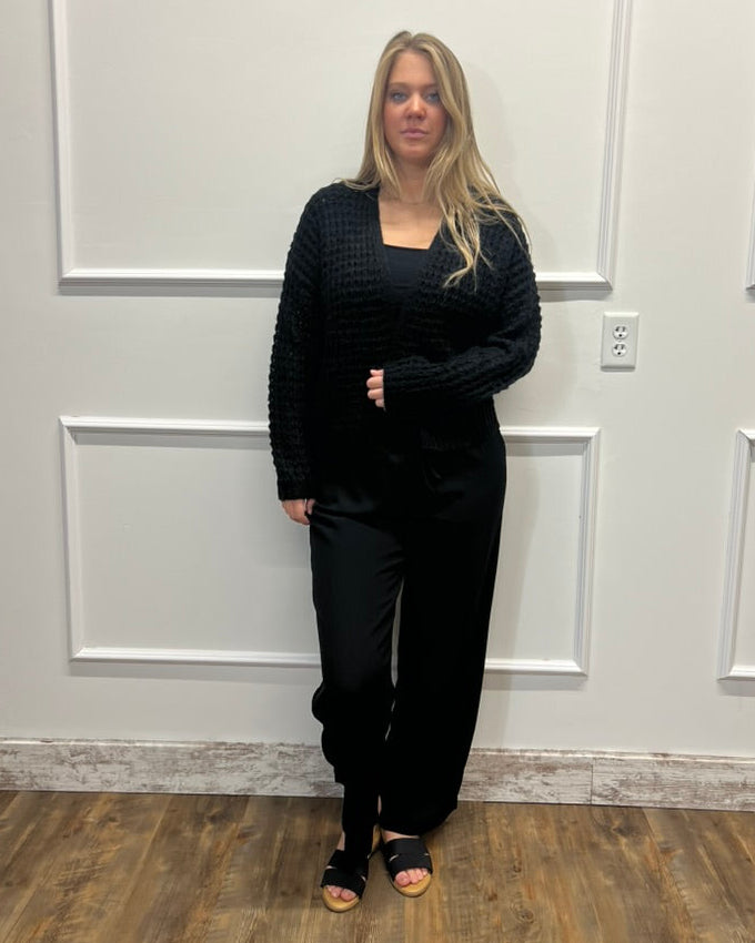 A woman stands indoors wearing a black textured knit long-sleeve top and black wide-leg pants, paired with black open-toe heels, against a white paneled wall with wood flooring.