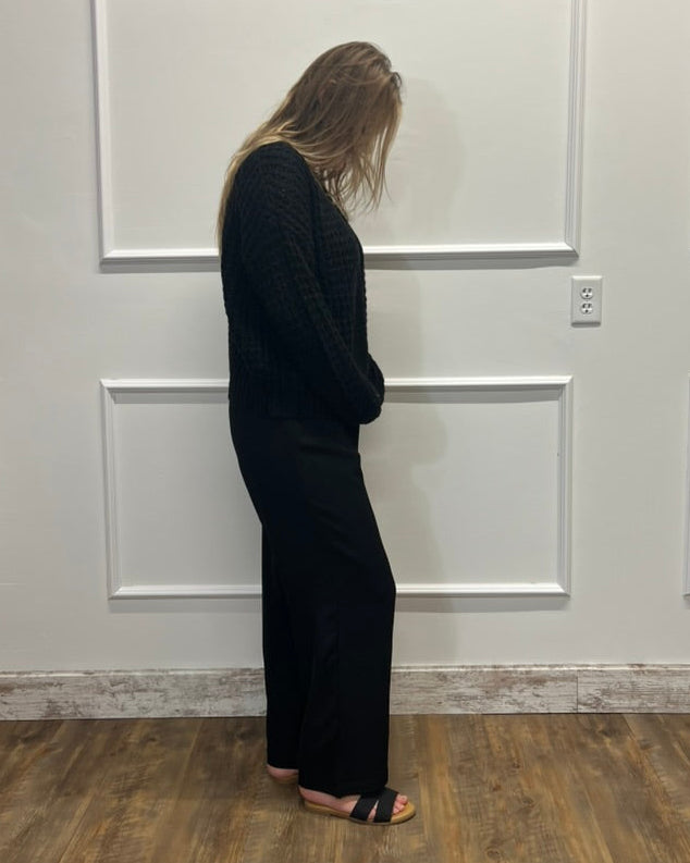 A woman stands in profile wearing a black textured knit long-sleeve top and matching black wide-leg pants, standing on a wood floor against a white paneled wall.