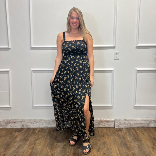 A woman modeling a black maxi dress with a yellow and white floral pattern, standing in front of a white paneled wall on a wood floor.