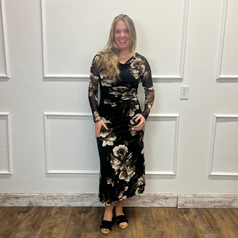 A woman modeling a long-sleeve black maxi dress with a beige and white floral pattern, standing in front of a white paneled wall on a wood floor.