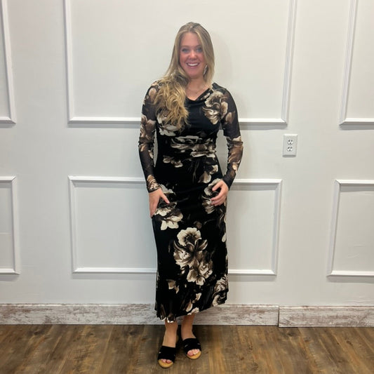 A woman modeling a long-sleeve black maxi dress with a beige and white floral pattern, standing in front of a white paneled wall on a wood floor.