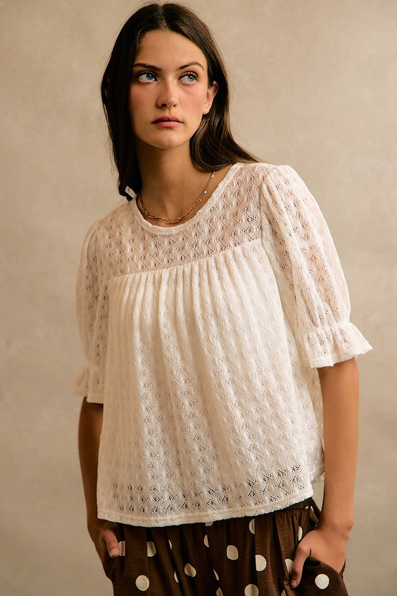 A woman wearing a cream-colored, semi-sheer eyelet lace short-sleeve blouse with a gathered bust, paired with brown polka-dot pants, standing against a neutral beige background.