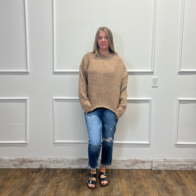 A woman stands in a room with white paneled walls and a wood floor, wearing a beige knit mock-neck sweater, blue distressed jeans, and black sandals.