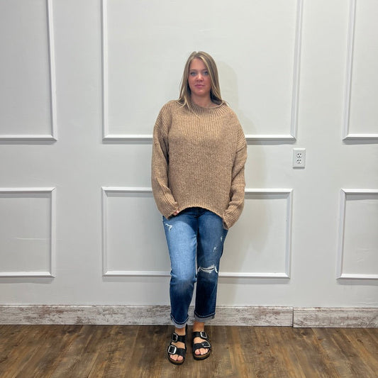 A woman stands in a room with white paneled walls and a wood floor, wearing a beige knit mock-neck sweater, blue distressed jeans, and black sandals.