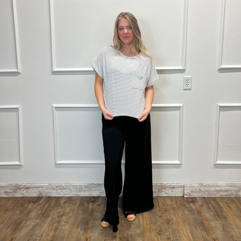 A woman stands in a room with white paneled walls and a wood floor, wearing a white short-sleeve top with thin black horizontal stripes and black wide-leg pants.