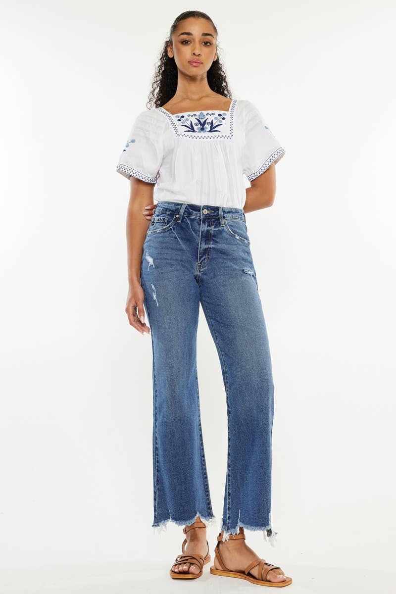A woman modeling a white short-sleeve top with blue floral embroidery on the chest and blue distressed wide-leg jeans with a raw hem, paired with brown leather sandals, against a plain white background.