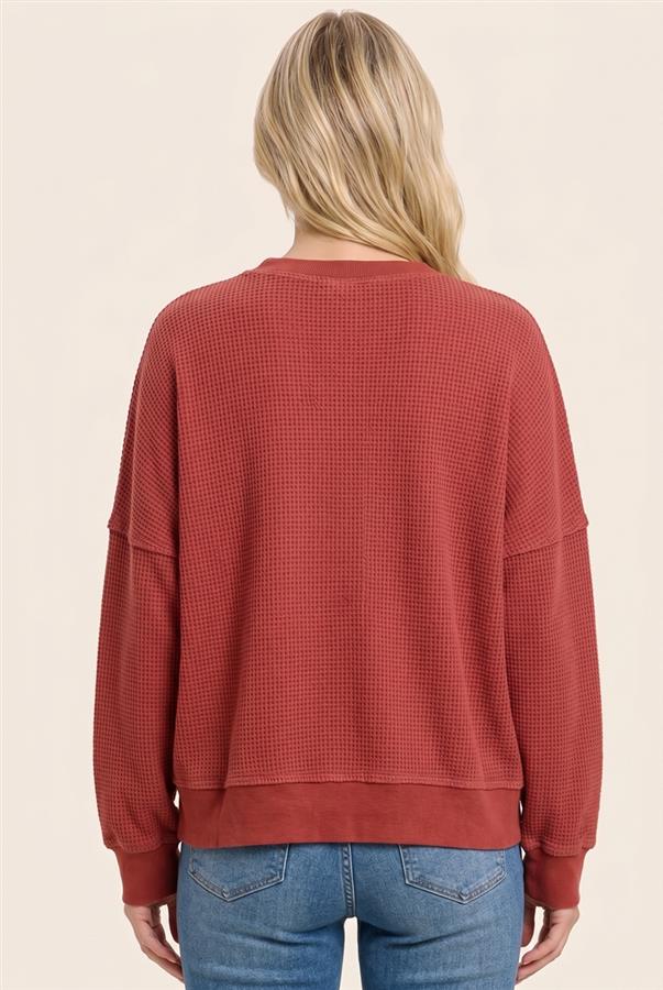 A woman viewed from the back wearing a textured, waffle-knit long-sleeve crewneck sweatshirt in a muted terracotta red, paired with blue jeans, against a solid light beige background.