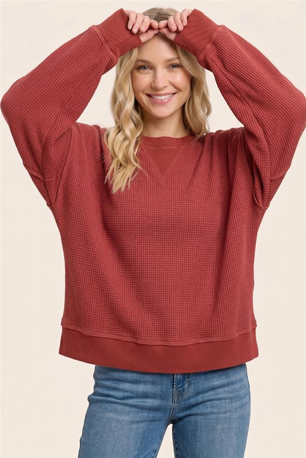 A woman wearing a rust-red waffle-knit crewneck sweatshirt and blue jeans, posing with her hands above her head against a light beige background.