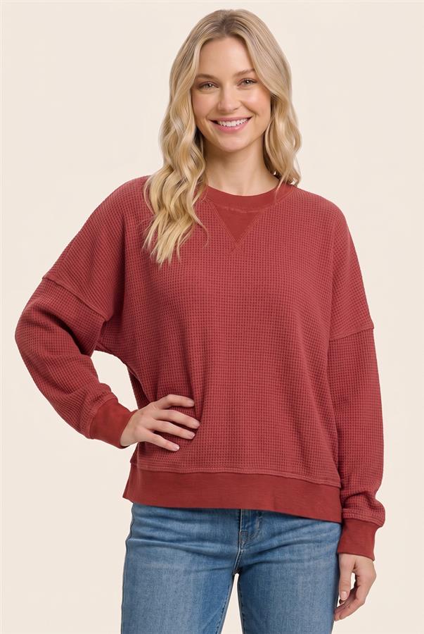 A woman wearing a rust-red waffle-knit crewneck sweatshirt and blue jeans, standing against a solid beige background.