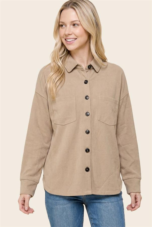 A woman wearing a beige button-up shirt jacket with two chest pockets and dark buttons, paired with blue jeans, against a solid light beige background.