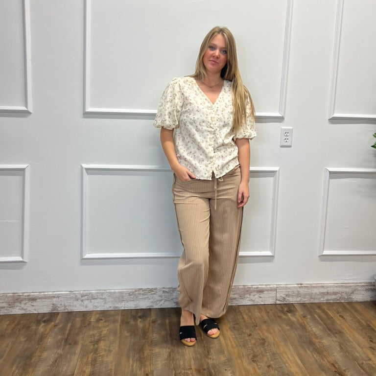 A woman stands against a white paneled wall on a wood floor, wearing a cream floral short-sleeve blouse and beige wide-leg pants with black slide sandals.