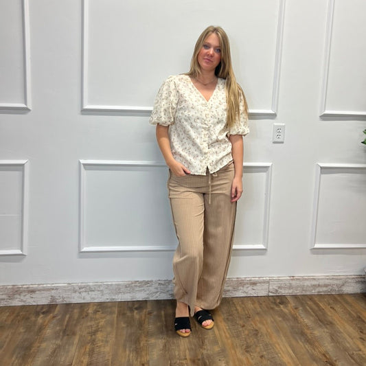 A woman stands against a white paneled wall on a wood floor, wearing a cream floral short-sleeve blouse and beige wide-leg pants with black slide sandals.