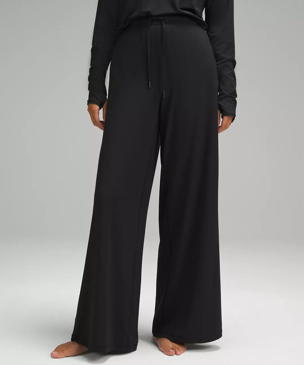 A model wearing black wide-leg drawstring pants and a matching black long-sleeve top, shown from the waist down against a neutral gray studio background.