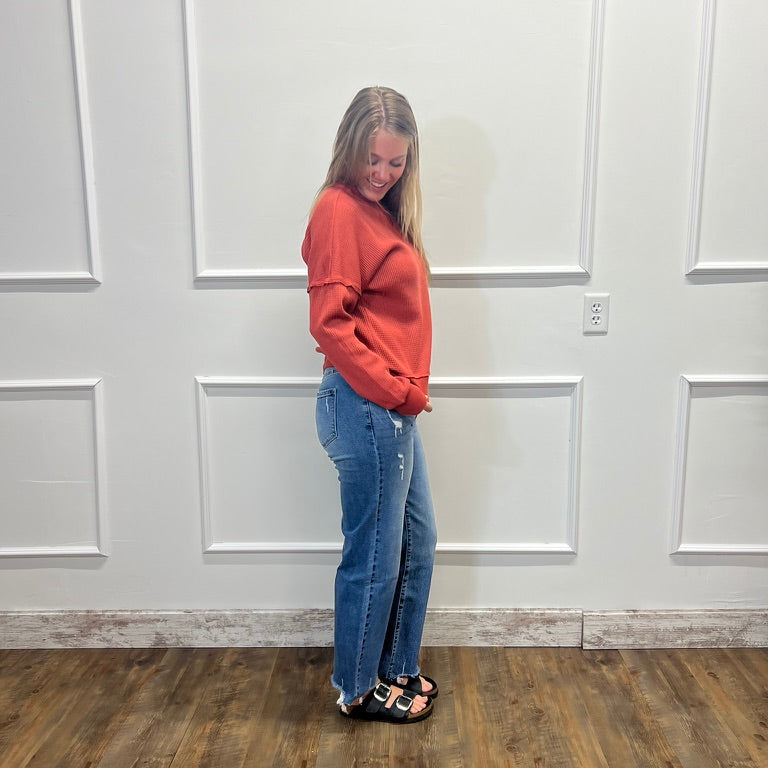 A woman stands in profile wearing a rust-orange long-sleeve sweatshirt, blue jeans, and black sandals, standing on a wood-look floor against a white paneled wall.