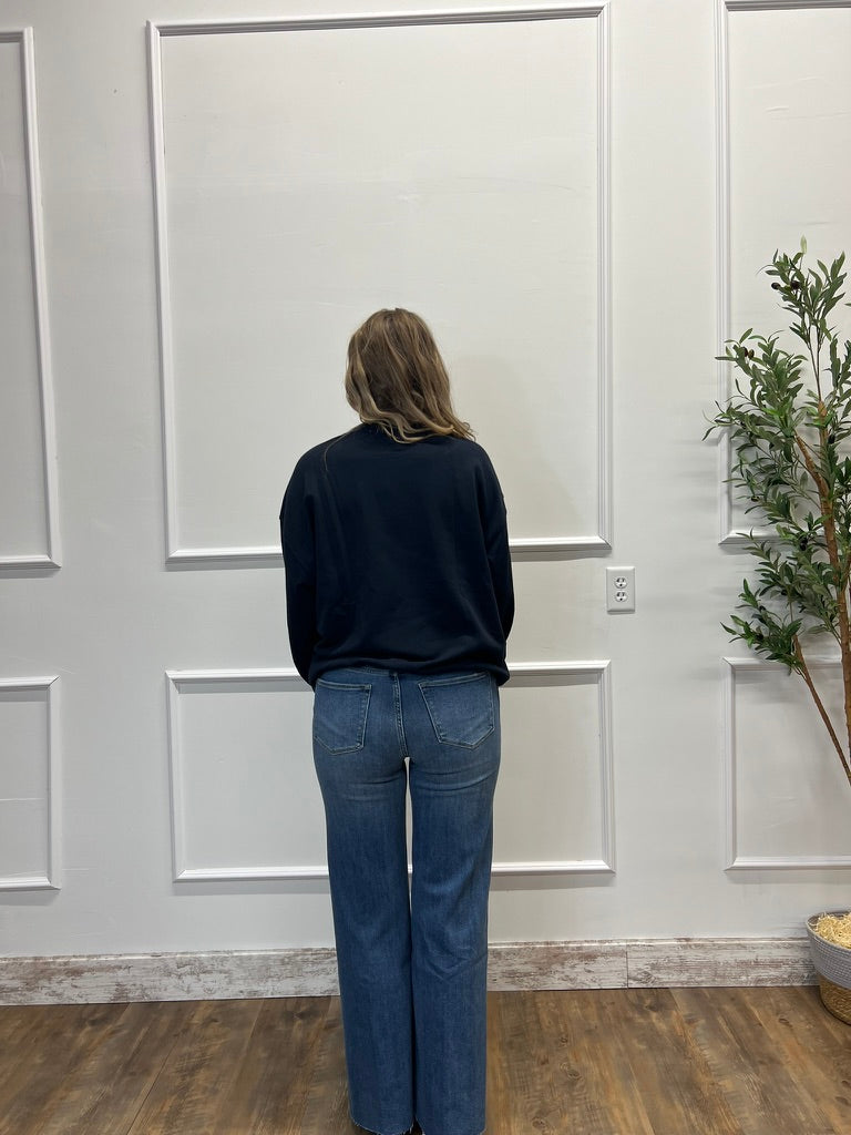 Person wearing a dark sweater and blue jeans standing in front of a white paneled wall.