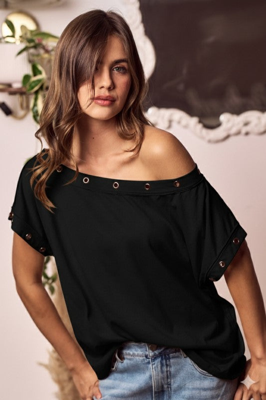 Woman wearing a black off-shoulder top with decorative eyelets, standing indoors.