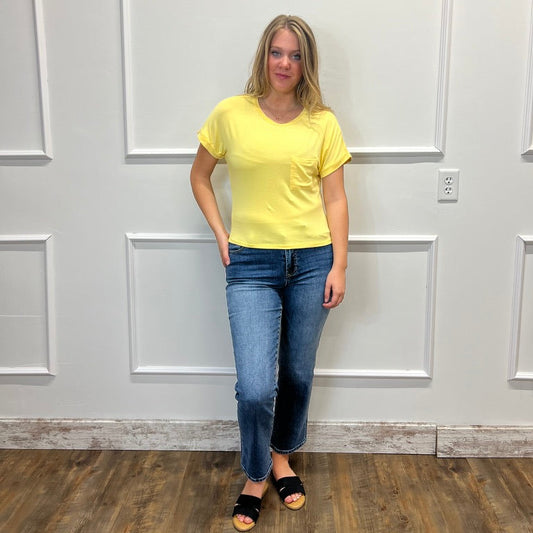 A woman stands indoors against a white paneled wall wearing a bright yellow short-sleeve crewneck t-shirt, blue jeans, and black slide sandals.