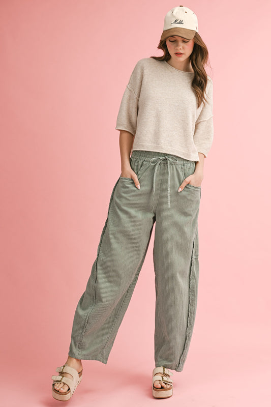 Woman wearing a beige sweater and green pants against a pink background