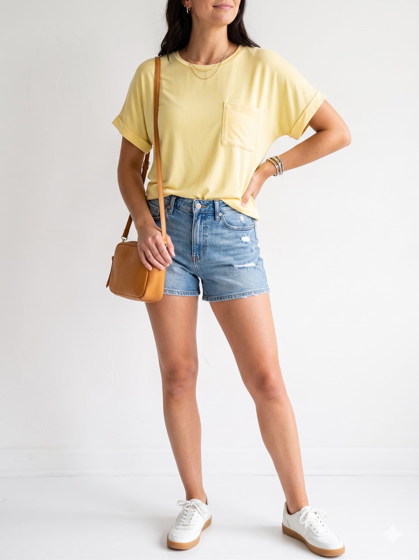 Woman wearing a yellow t-shirt, denim shorts, and white sneakers with a brown crossbody bag on a light background.