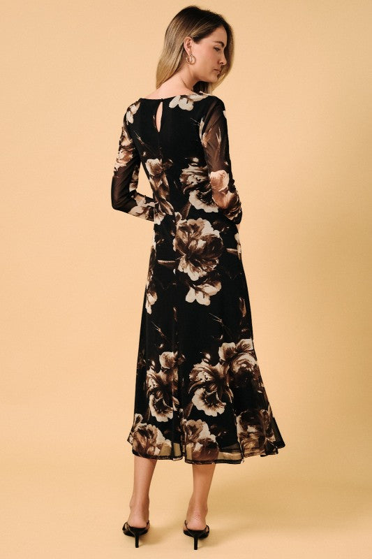 A woman modeling a long-sleeve midi dress with a black base and a large floral print in beige and brown, shown from the back against a solid beige studio background.