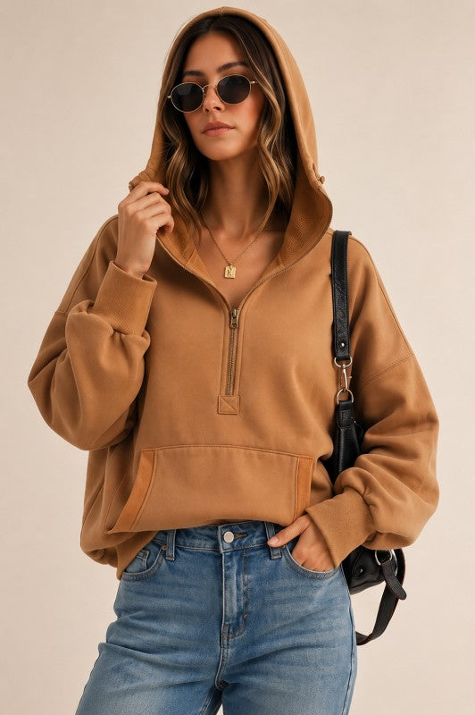 A woman wearing a camel brown half-zip hoodie with a kangaroo pocket, paired with blue jeans and a black shoulder bag, photographed against a solid beige background.