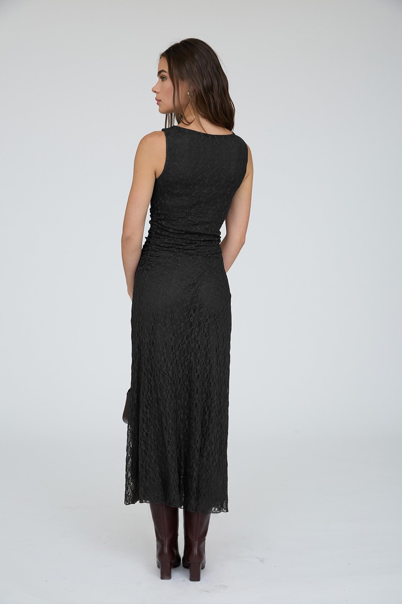 A woman shown from the back wearing a sleeveless black lace midi dress and dark brown knee-high boots against a plain light gray studio background.