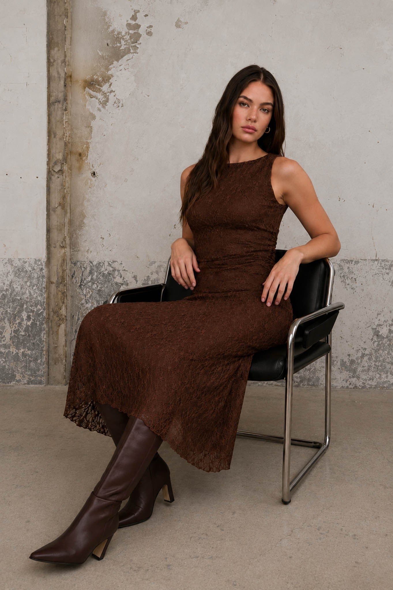 Woman in a brown dress and boots sitting on a black chair against a textured wall.