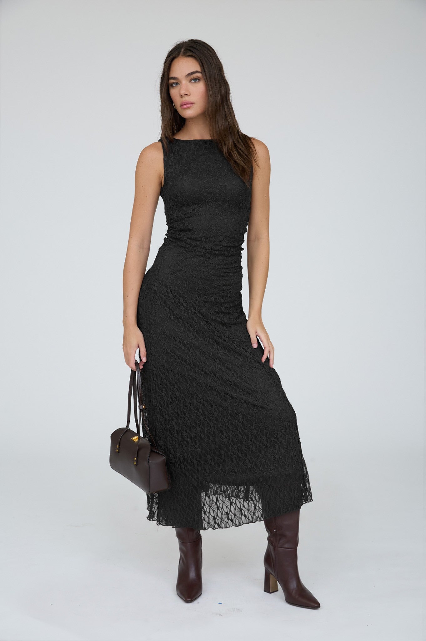 A woman modeling a sleeveless black midi dress with a textured, ribbed knit pattern and a sheer lace hem, styled with dark brown leather knee-high boots and a dark brown handbag against a light gray studio background.