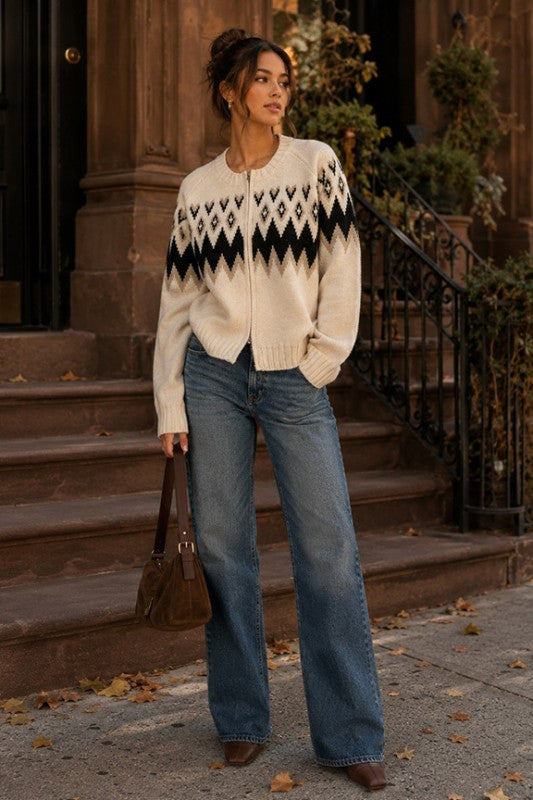 A woman stands on outdoor stone steps wearing a cream knit zip-front sweater with a black geometric pattern across the chest, blue wide-leg jeans, and brown ankle boots, while holding a brown handbag.