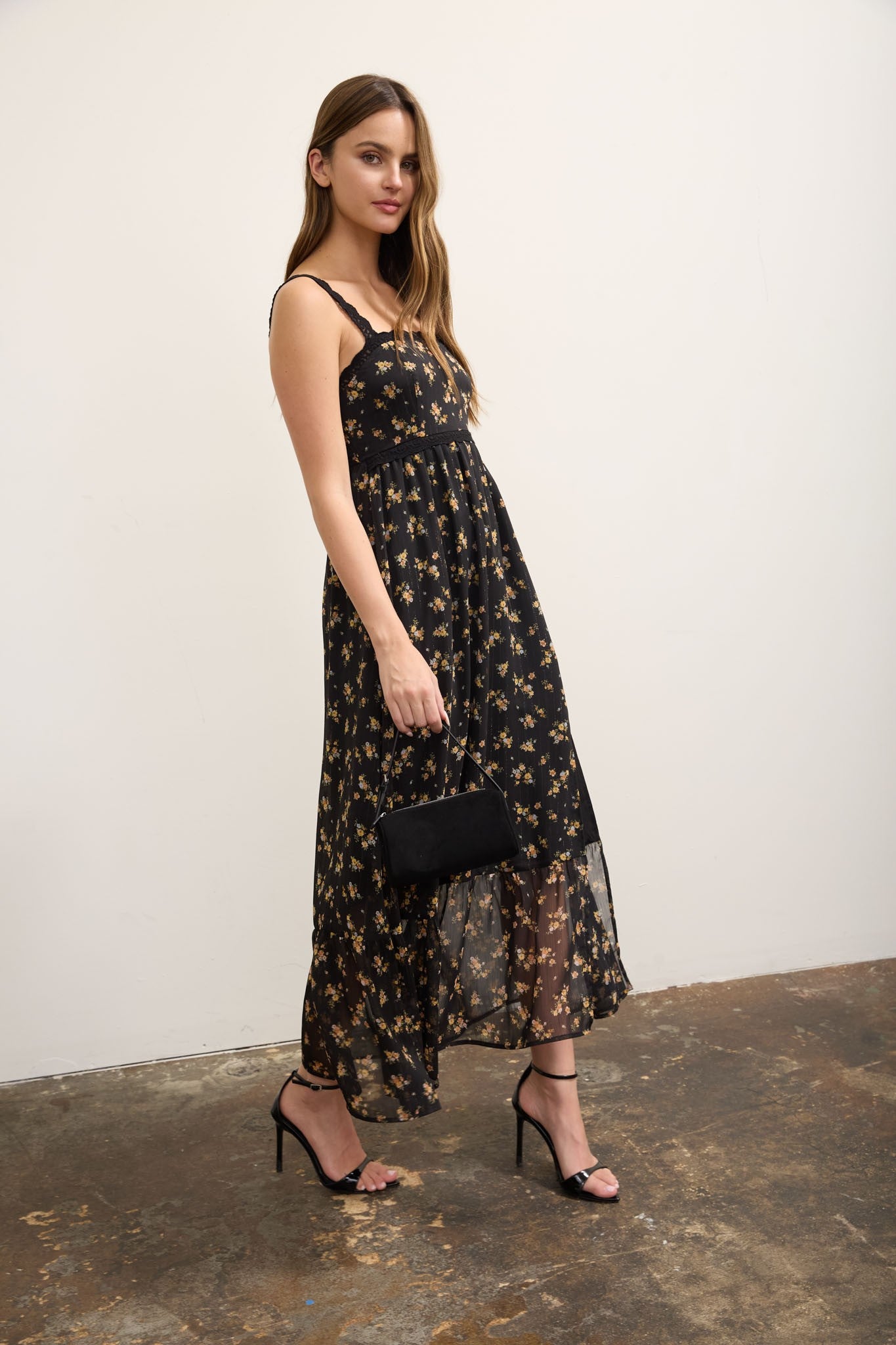 A woman modeling a black midi dress with a beige floral pattern, styled with black strappy heels and holding a small black clutch bag, set against a plain off-white wall and textured concrete floor.
