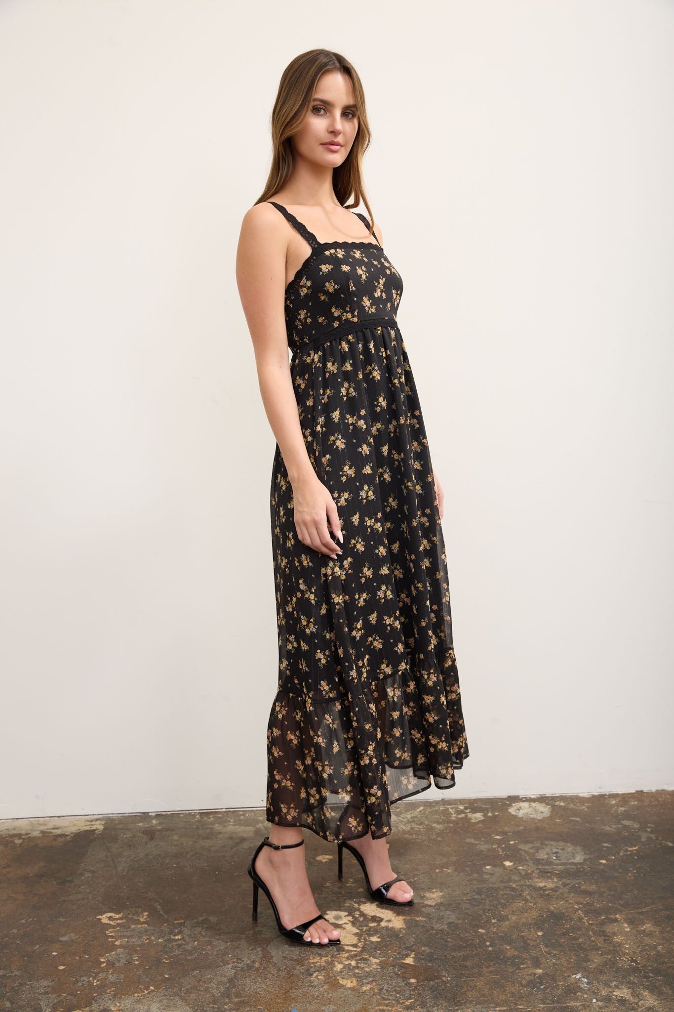 A woman modeling a black midi dress with a gold floral pattern, featuring thin shoulder straps and a square neckline, paired with black high-heeled sandals against a neutral studio backdrop.
