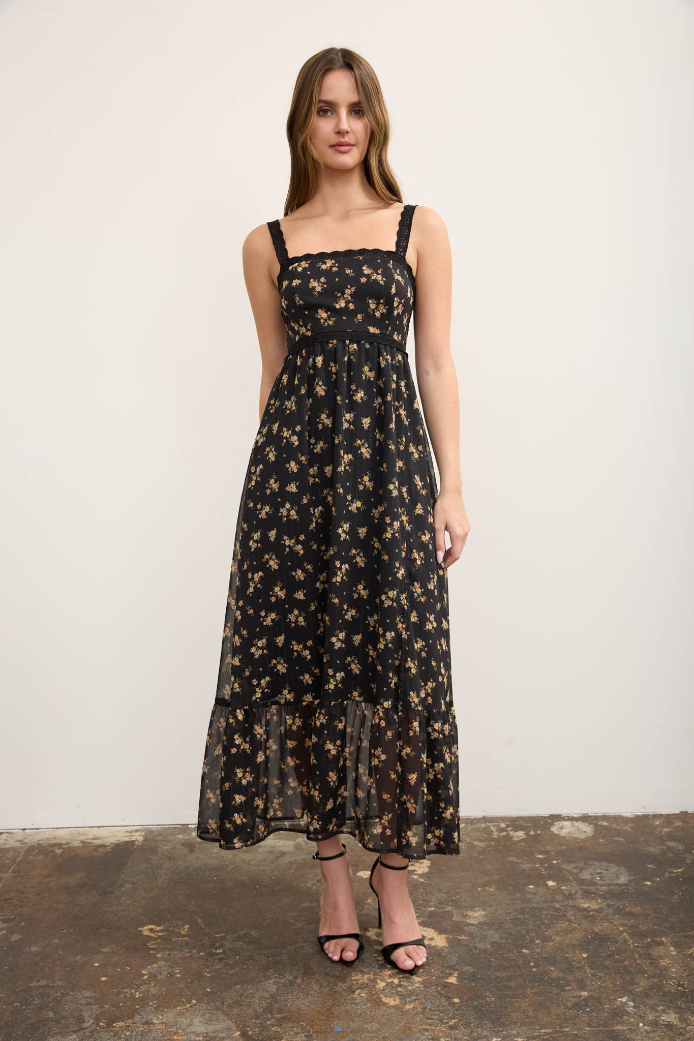 A woman modeling a black midi dress with a beige floral pattern, featuring thin shoulder straps and a tiered ruffle hem, standing against a plain off-white wall.