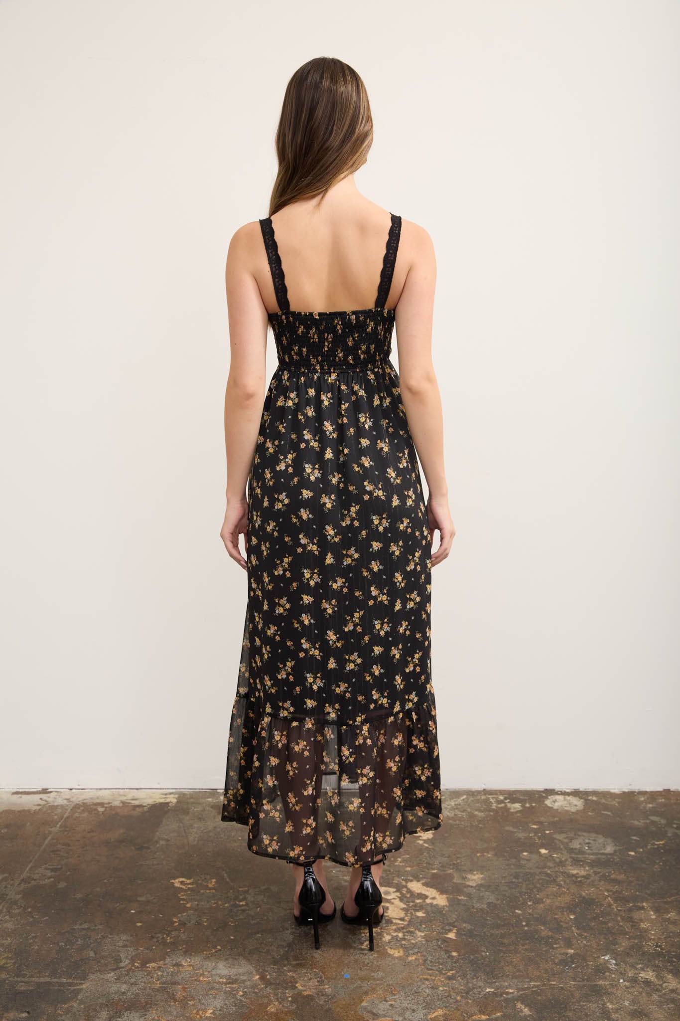 A woman viewed from behind wearing a black spaghetti-strap maxi dress with a small beige and brown floral print, standing against a plain off-white wall.