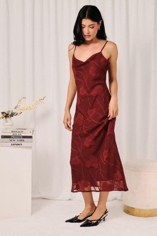 A woman models a burgundy midi slip dress with a tonal floral jacquard pattern, styled with black pointed-toe heels against a white curtain backdrop.