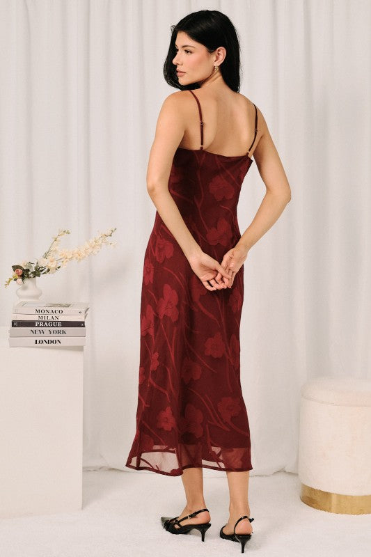 A woman modeling a burgundy midi slip dress with a tonal floral jacquard pattern, shown from the back against a white curtain backdrop.