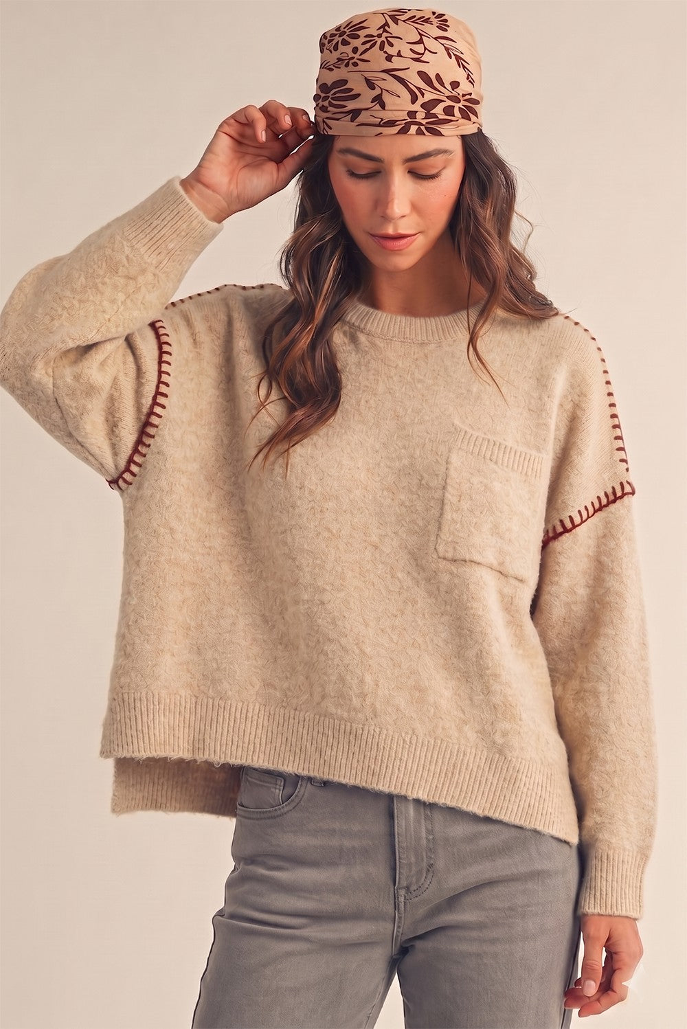 A woman wearing a beige knit sweater with brown decorative stitching on the sleeves, paired with gray jeans and a brown patterned beanie, against a plain beige background.