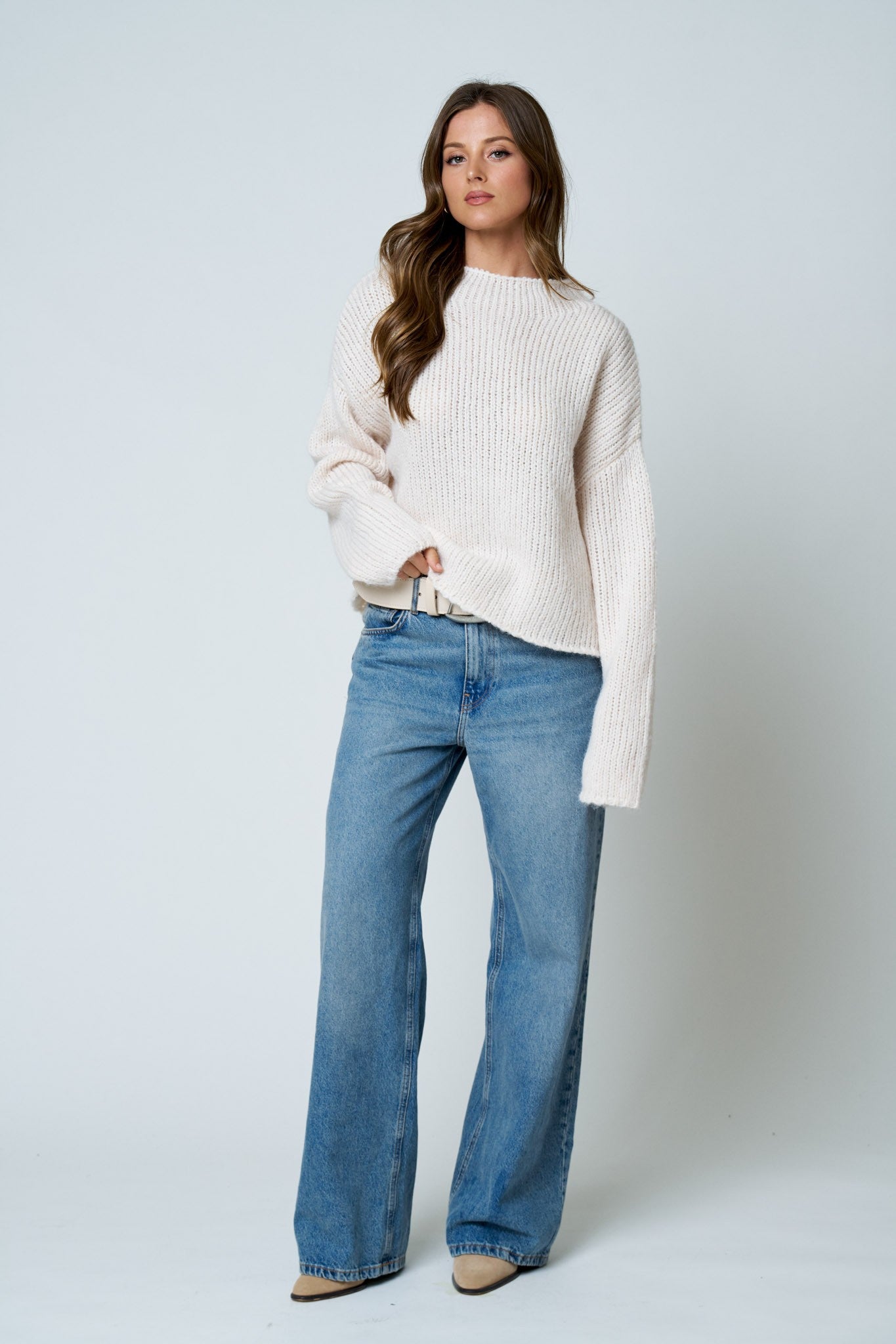 A woman modeling a cream ribbed knit sweater and wide-leg blue jeans against a light gray studio background.