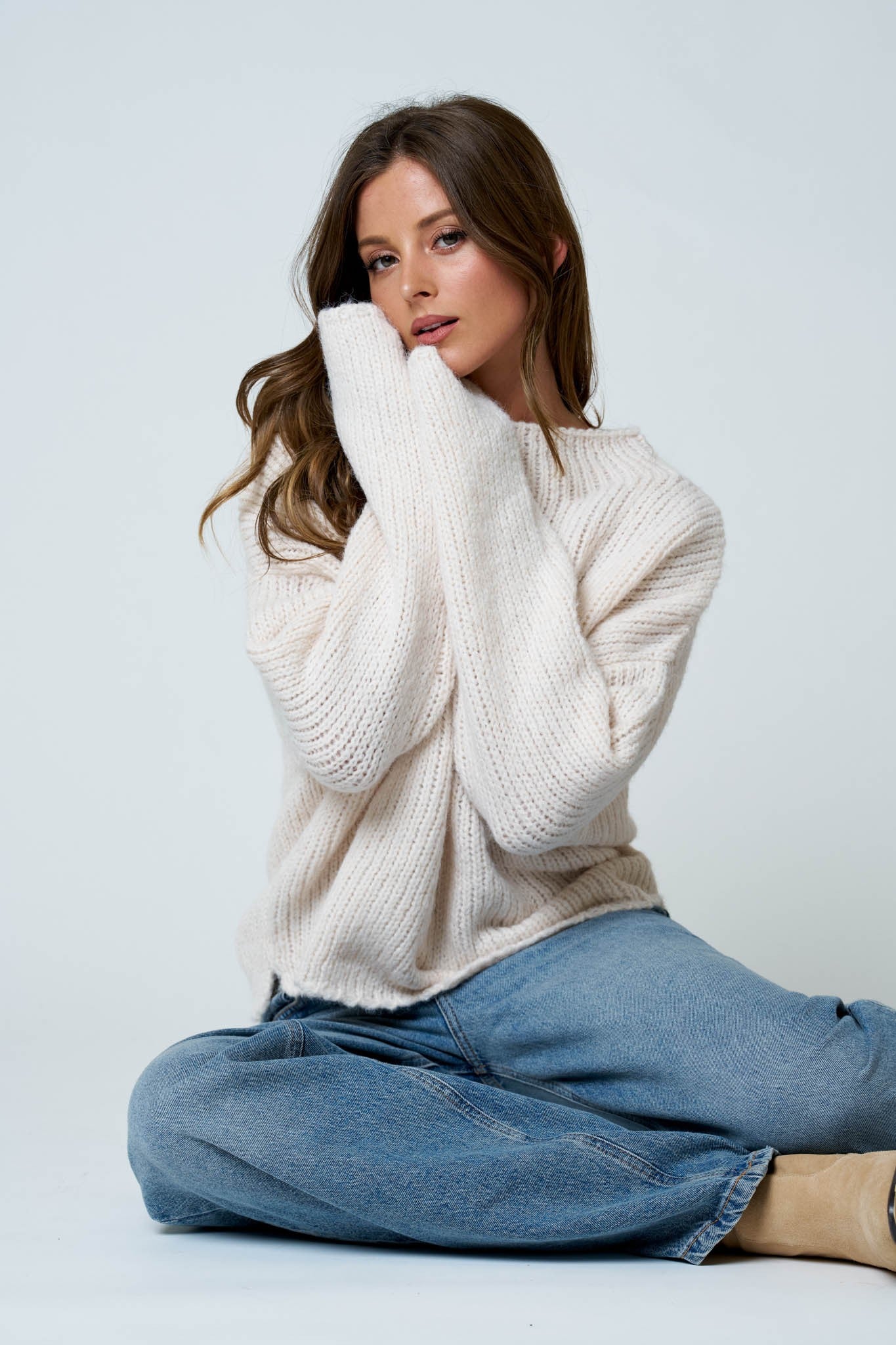 Woman wearing a white sweater and blue jeans sitting on a light gray background