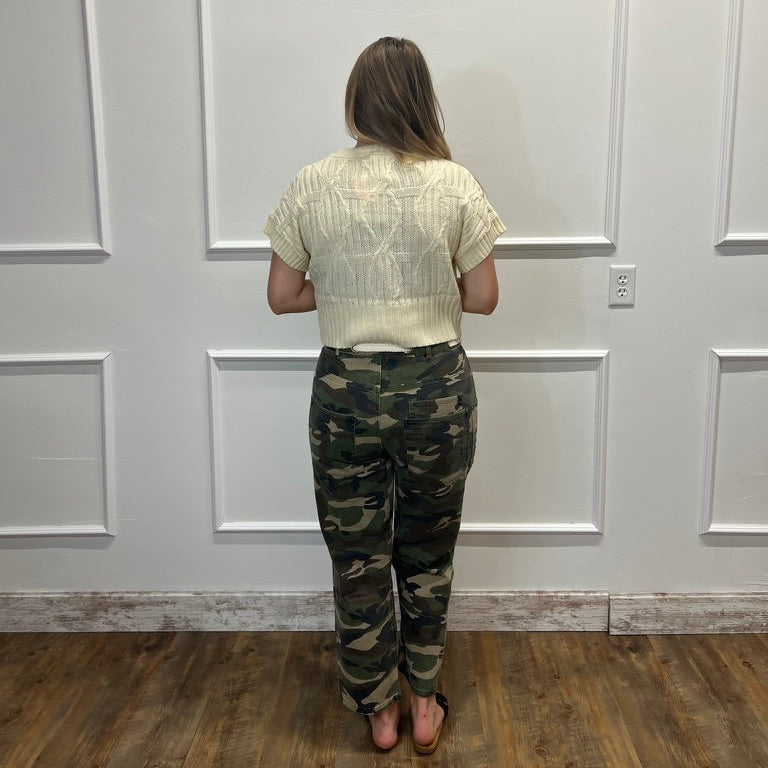 A woman stands with her back to the camera wearing a cream-colored textured short-sleeve crop top and camouflage pants, standing in front of a white paneled wall on a wooden floor.