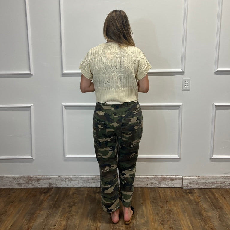 A woman stands with her back to the camera wearing a cream-colored textured short-sleeve crop top and camouflage pants, standing in front of a white paneled wall on a wooden floor.