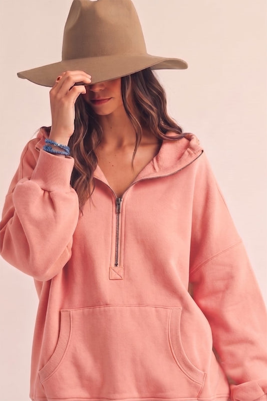 A woman wearing a dusty pink half-zip hoodie and a wide-brimmed tan hat, posing against a solid pale pink background.