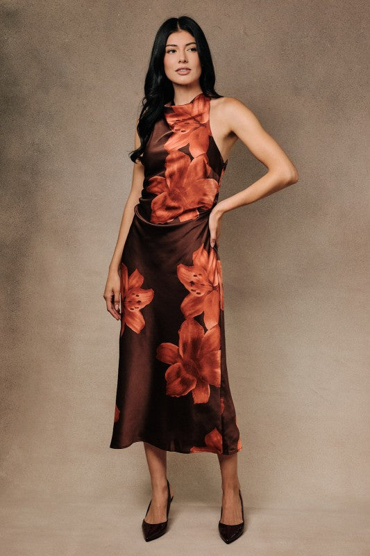 A woman modeling a sleeveless high-neck midi dress with a brown satin-like base and large rust-orange floral print, posed against a textured warm beige studio backdrop.