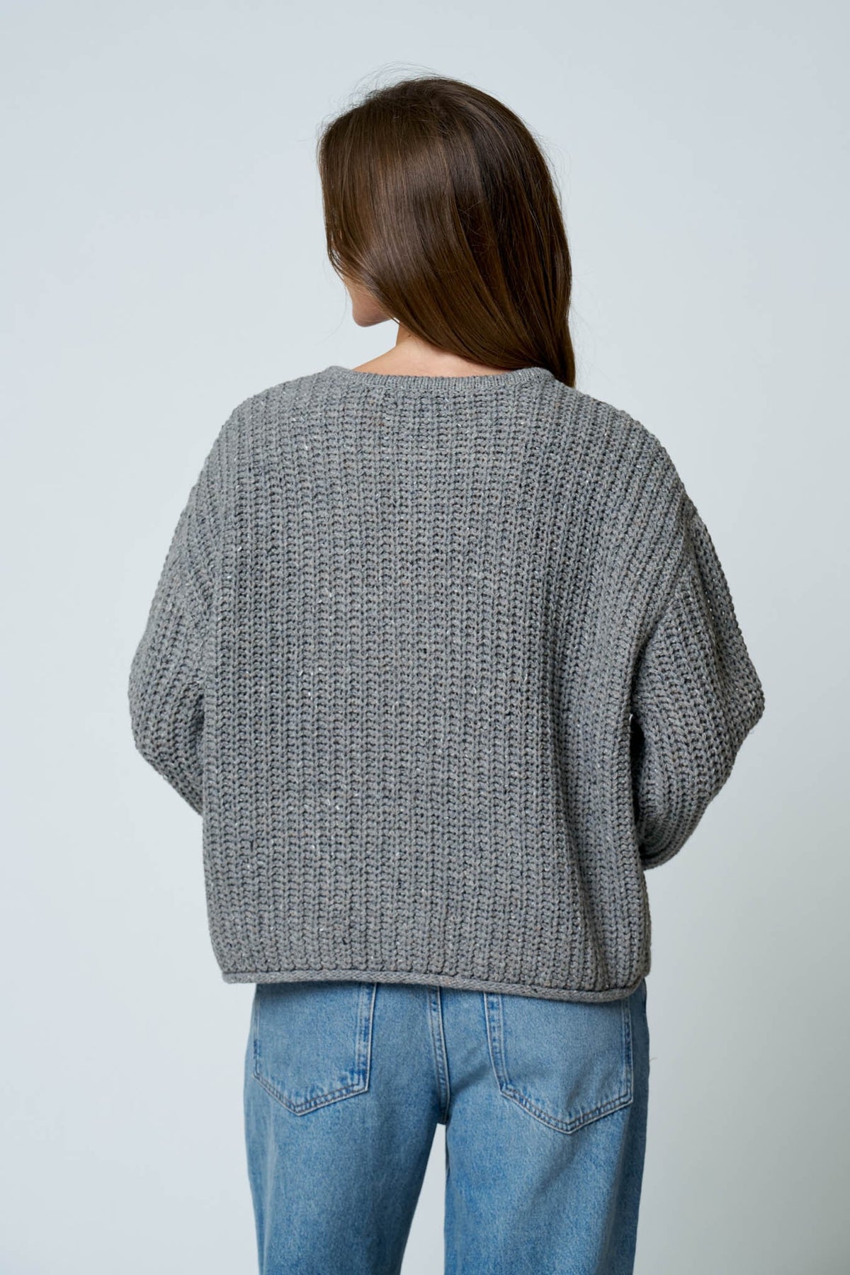 A woman viewed from behind wearing a gray ribbed knit sweater and blue jeans against a plain light gray background.