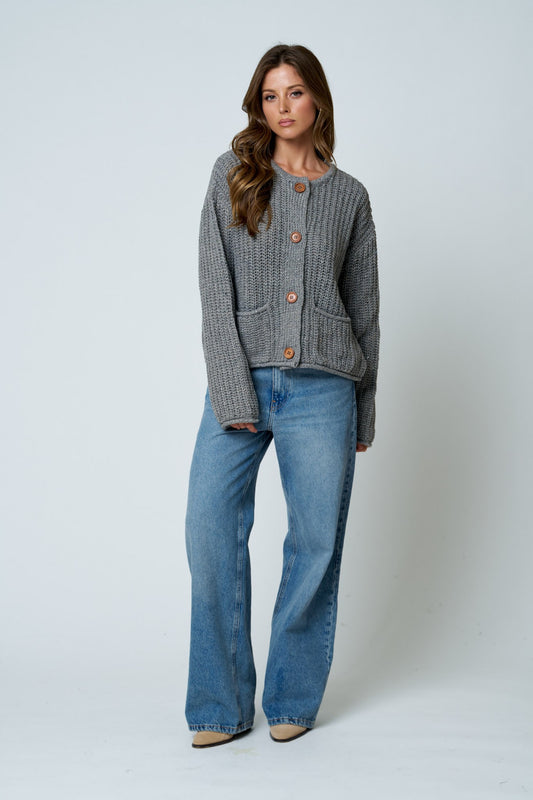 A woman modeling a gray ribbed knit cardigan with brown buttons, paired with wide-leg blue jeans, standing against a light gray studio background.
