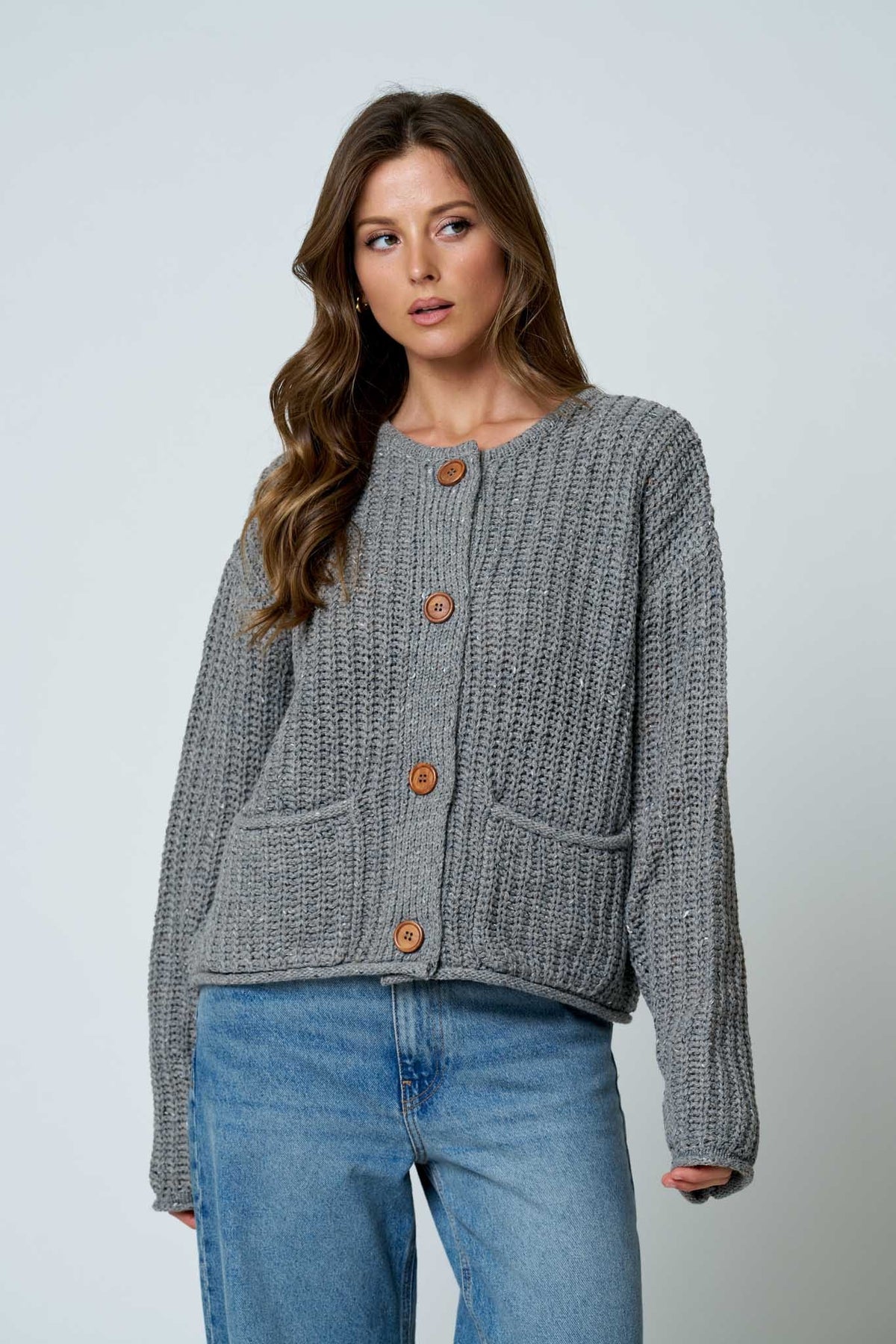 A woman wearing a gray ribbed knit cardigan with brown buttons and blue jeans, standing against a light gray studio background.