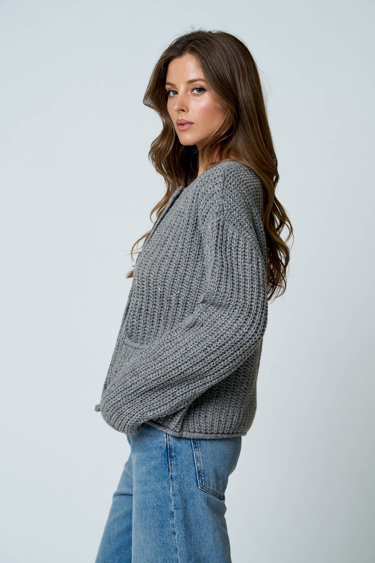 A woman wearing a chunky gray cable-knit cardigan sweater and blue jeans, photographed against a plain light gray studio background.