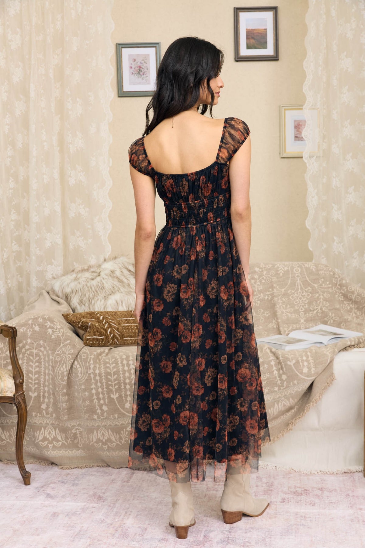 A woman stands with her back to the camera wearing a dark floral midi dress with sheer cap sleeves and an open back, paired with beige ankle boots, in a vintage-style living room setting.